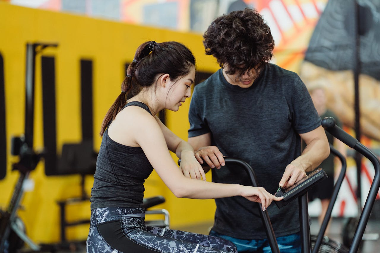 hero-img-02 A couple in activewear setting up an elliptical trainer during a workout session in a modern gym.